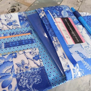 May include: Open blue and white fabric wallet with floral and polka dot patterns. The wallet has multiple pockets, a clear sleeve, and a blue ribbon. A blue pen and a business card are visible.
