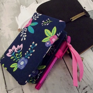 May include: A navy blue fabric pouch with a floral pattern in pink, blue, green, and white. The pouch is folded and has a pink ribbon attached to it. A pink pen is sticking out of the pouch.