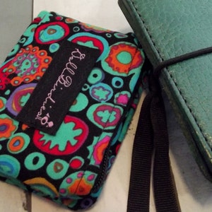 May include: A colorful, patterned wallet with a black background, teal, orange, and pink circular designs. The wallet has a black label with the text "Ball Birdies" in pink. Next to it is a teal leather notebook with a black elastic band.