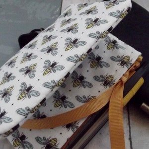May include: A fabric-covered journal with a bee pattern. The cover is white with a repeating pattern of bees in yellow, black, and gray. An orange ribbon is tied around the journal. A black case is visible in the background.