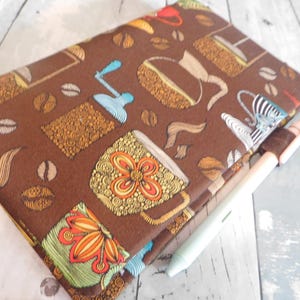 May include: A brown fabric notebook cover featuring a repeating pattern of coffee beans, mugs, and coffee grinders in shades of orange, yellow, green, and blue. A light-colored pen is tucked into the fabric pen loop on the side of the cover.