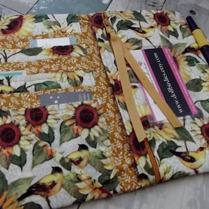 May include: A fabric wallet with a sunflower and bird pattern. The wallet has multiple pockets and a zipper closure. A pen and a ribbon are visible. The website address www.dollbirdies.etsy.com is printed on a card.