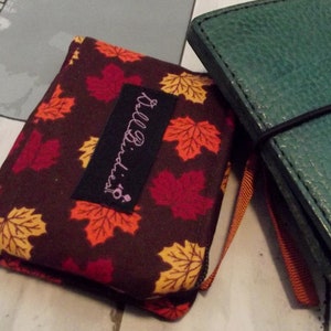 May include: A brown fabric journal cover with a fall leaf pattern in red, orange, and yellow. The cover has a black label with the text "Kill Birdies!" in white script. A green leather journal is next to it.