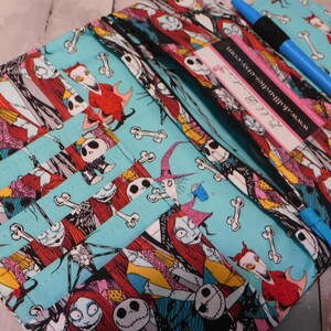 May include: A zippered pencil case featuring a colorful print of characters from The Nightmare Before Christmas on a light blue background. The case has multiple compartments and a blue zipper pull. A blue pen and a label with the website www.dollbirdies.etsy.com are visible.