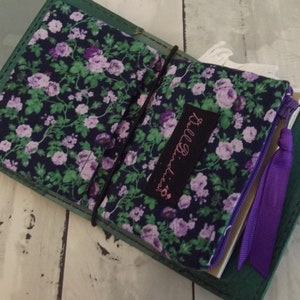May include: A dark green leather journal cover featuring an interior fabric lining with a purple and green floral pattern. A black fabric label with the text Doll Birdies is stitched onto the fabric, and a purple ribbon pull is attached to the zipper.