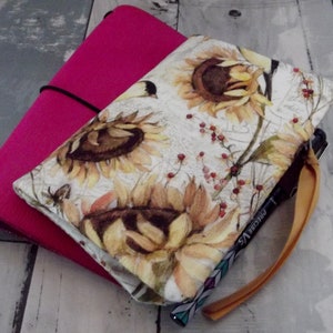 May include: A fabric journal cover featuring a sunflower print in yellow, brown, and cream, layered over a solid magenta notebook. A black elastic band secures the cover, and a black Precise V5 pen with a yellow ribbon bookmark is attached.