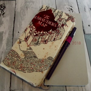 May include: A cream-colored notebook cover with a map design featuring a red castle and the text "The Marauder's Map". The cover is slightly worn and has a vintage look. The notebook cover is lying on top of a gray notebook with the year "2018" printed on the front.