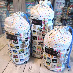 May include: Three cylindrical drawstring bags of varying heights featuring a white fabric printed with colorful vintage-style cameras and small hearts. Each bag has a blue drawstring cord, a toggle closure, and a black label reading Doll Birdies.