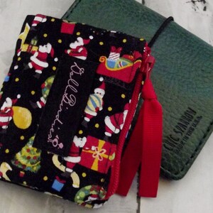 May include: A black zippered pouch with a Christmas-themed pattern featuring Santa Claus, gifts, and Christmas trees. A red ribbon is attached to the zipper. A green leather notebook is next to the pouch. The pouch has a tag that says "Ball Birdies".