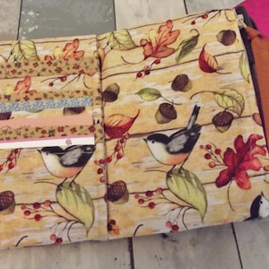 May include: A fabric zippered pouch with a fall-themed design. The pouch features a beige background with brown branches, red berries, green leaves, and brown acorns. Two small birds with black heads and white bellies are perched on branches. The pouch has multiple pockets on the inside.