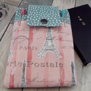 May include: A pink and white fabric case with a Parisian theme, featuring the Eiffel Tower and the words "Paris" and "Carte Postale." The case has a teal flap with white polka dots and a snap closure. Two pens are visible in the pockets.