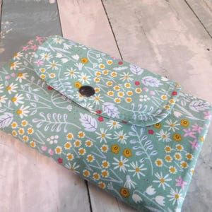 May include: A floral print wallet with a button closure. The wallet features a light green background with a pattern of white daisies, yellow centers, and pink accents. The wallet is closed.