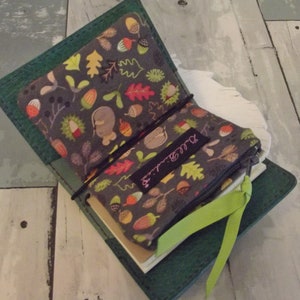 May include: Open green leather journal with a patterned fabric interior featuring acorns, leaves, and a squirrel. A matching zippered pouch with the text "Bell Bindings" is inside. A green ribbon bookmark is visible.