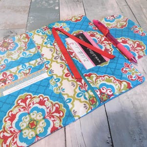 May include: A fabric organizer featuring a bright blue, red, and green floral pattern. It includes multiple interior pockets, a red ribbon bookmark, and a pink pen. A business card with the text www.dollbirdie.com is tucked into a slot.