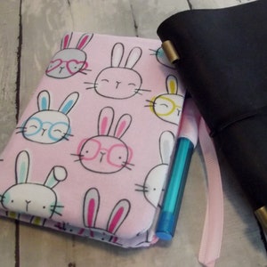 May include: A pink notebook with a bunny rabbit pattern. The notebook has a blue pen tucked inside and is closed. The notebook is sitting on a white wooden surface.