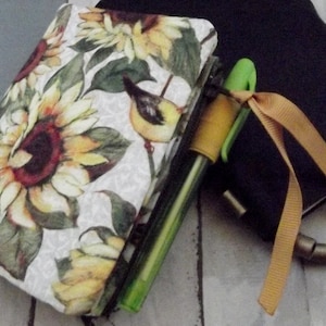 May include: A white fabric notebook cover with a sunflower and bird design. The cover is attached to a black notebook with a brown ribbon.