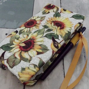 May include: A fabric notebook cover featuring a sunflower and bird pattern in yellow, red, and green on a cream background. A pen and a yellow ribbon are visible.