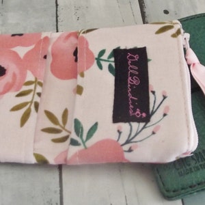 May include: A floral print wallet with a zipper closure and a pink ribbon pull. The wallet features a pattern of pink flowers and green leaves on a light background. A green leather notebook is next to the wallet.