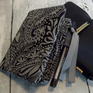 May include: A black and gray paisley patterned fabric pouch with a zipper closure. A black pen with a silver clip is inside the pouch. The pouch is lying on a white wooden surface.
