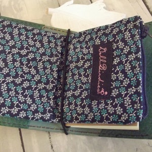 May include: A green fabric-covered notebook with a zippered pocket and a blue ribbon. The pocket has a floral pattern and a purple label that says "Bulla Buddies".