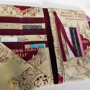 May include: A fabric wallet with a map design, featuring burgundy accents and multiple pockets for cards and cash. The wallet has a zipper closure and a burgundy ribbon. A purple pen rests on the side.