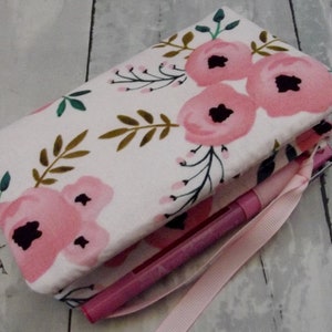 May include: A floral print pencil case with a pink ribbon. The case is white with pink flowers and green and brown leaves. A pink pen is visible inside the case.