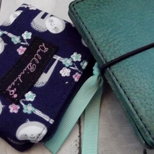 May include: Two small notebooks, one with a blue fabric cover with a sloth pattern and pink flowers, and the other with a teal leather cover. The notebook with the sloth pattern has a black label that says "Dream Big". The teal notebook has a black elastic band and the text "Chic & Spark Made in Italy" on the cover.