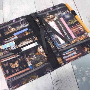 May include: Open, black fabric wallet with a detailed print of a cabinet of curiosities, featuring skulls, books, butterflies, and other vintage items. The wallet has multiple compartments, a zipper closure, and a black strap. An orange pen rests on the side.