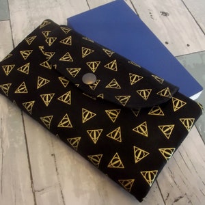 May include: A black fabric wallet with a gold repeating pattern of the Deathly Hallows symbol. The wallet has a flap with a button closure.