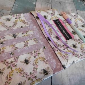 May include: A fabric planner cover featuring a cream background with a pattern of bees and floral wreaths in pink and yellow. The interior includes multiple card slots, a lavender ribbon bookmark, and a pen holder with a light pink and blue pen.