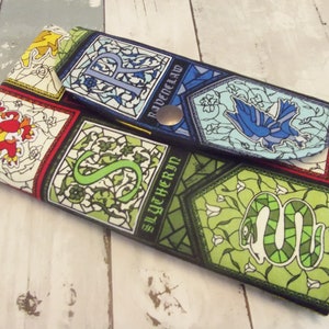 May include: A fabric wallet featuring stained-glass style designs with the Hogwarts houses' emblems. The wallet has a snap closure and includes the Ravenclaw and Slytherin house crests. The color palette includes green, blue, red, and yellow.