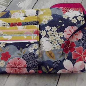 May include: A blue and gray fabric wallet with a floral and bunny pattern. The wallet has a pink flap and a black ribbon.