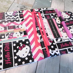 May include: A fabric wallet featuring a black and white Hello Kitty pattern with pink chevron accents. The interior includes multiple card slots, a zippered compartment, and a clear plastic ID window. A pink pen rests on the open wallet.
