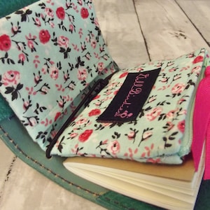 May include: A teal leather notebook cover with a floral fabric insert and a pink ribbon. The fabric has a light blue background with pink roses and black leaves. The cover has a small black label with the text "Lil' Bits" on it.