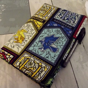 May include: A fabric book cover featuring stained-glass style panels with the names and emblems of Hogwarts houses: Hufflepuff, Ravenclaw, and others. The cover is in shades of yellow, blue, and green, with a black pen tucked into the side.