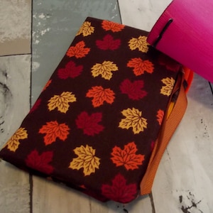 May include: A brown fabric pouch with a zipper closure and an orange ribbon. The pouch is decorated with a pattern of colorful maple leaves.