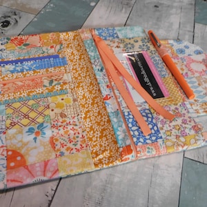 May include: Open patchwork wallet with a floral pattern in shades of orange, yellow, blue, and pink. The wallet has multiple card slots, a zippered compartment, and an attached coin purse. An orange pen rests on the wallet, and a business card is visible.