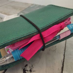May include: A teal leather journal with a black elastic band, holding a pink ribbon, a pink zippered pouch, and a blue pen. The pen has the text "Z-Grip" on it.