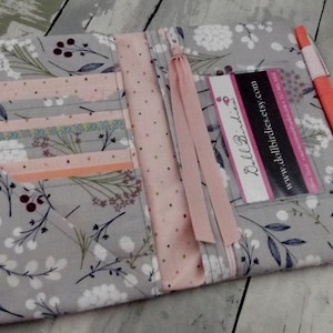 May include: Open gray fabric wallet with floral and berry pattern. Interior features card slots, a pink ribbon, and a business card. An orange pen rests on the side. The wallet is designed for organizing cards and cash.