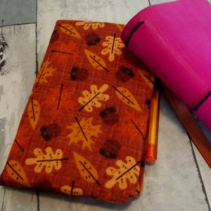 May include: A fall-themed notebook cover with a pattern of orange and yellow leaves and acorns on a rust-colored background. A bright pink notebook and a pen are also visible.