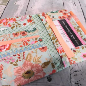 May include: Open floral print fabric wallet with a peach-colored ribbon and a matching pen. The wallet features a light green background with pink, orange, and red flowers. The text "www.dollbirdies.etsy.com" is visible on a label.
