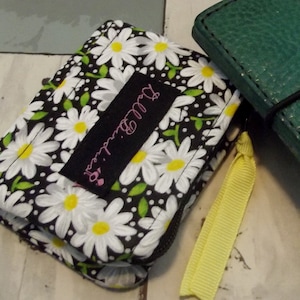 May include: A black fabric wallet with a white daisy pattern and a black label that says "Dutchess" in pink lettering. The wallet has a yellow zipper pull and a green leather wallet in the background.