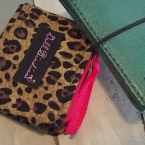 May include: A small leopard print wallet with a black label that says "Darling." The wallet has a pink zipper pull and is closed. The wallet is sitting on a white surface next to a green leather notebook with a black elastic band.