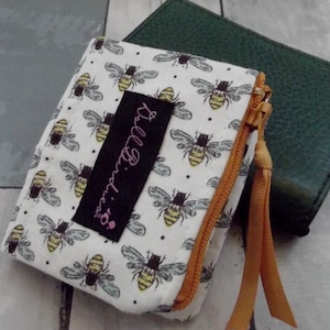 May include: A white zippered pouch with a bee print and a black label that says "Dollybird's". The pouch has a gold zipper and a brown zipper pull.