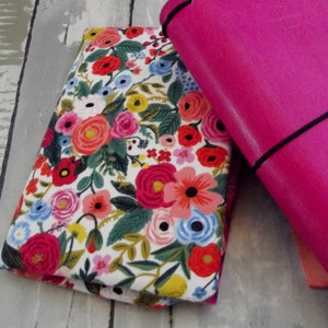 May include: A white fabric with a floral pattern in shades of pink, red, blue, and green. The fabric is folded and placed on top of a bright pink notebook.