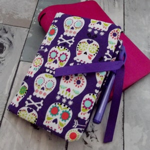 May include: Purple fabric notebook cover with a colorful sugar skull pattern. The cover has a purple ribbon and a purple pen.