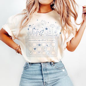 May include: Cream-colored t-shirt with a blue floral design and the text "communication looks different for everyone." The design includes a keyboard-like graphic with various icons. The shirt is made of a soft material.