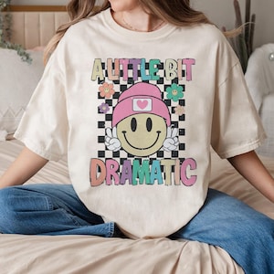May include: Cream-colored t-shirt with the text "A Little Bit Dramatic" in colorful, retro-style lettering. The graphic includes a smiley face wearing a pink beanie and making a peace sign, set against a black and white checkered background.