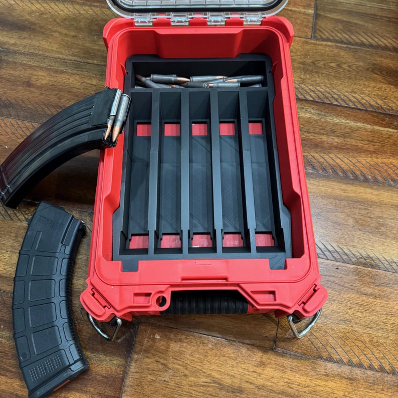 May include: A red storage case with a black interior, designed to hold ammunition. The case is open, revealing several compartments and a magazine. Two black magazines are placed next to the case, with bullets visible.