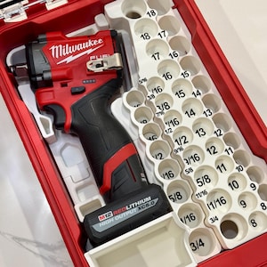 May include: A red and black Milwaukee FUEL impact driver with a M12 REDLITHIUM HIGH OUTPUT XC5.0 battery. The driver is in a red and white case with a white plastic insert that holds a variety of socket sizes. The socket sizes are labeled in fractions of an inch, from 3/4 inch to 5/16 inch.